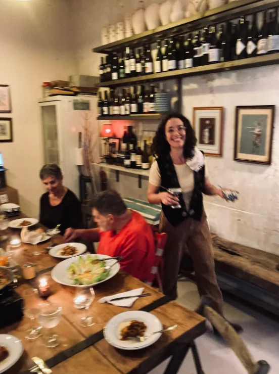 Gaby sonriendo junto a una mesa de cena con los platos servidos en Jugo, con la estantería de vinos detrás